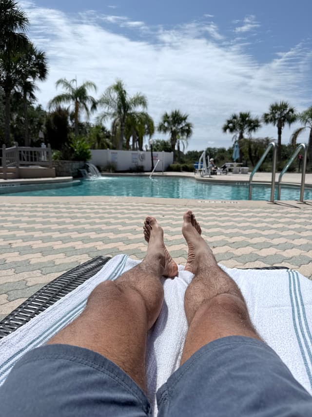 A person's legs resting on a lounge chair by a swimming pool under a sunny sky